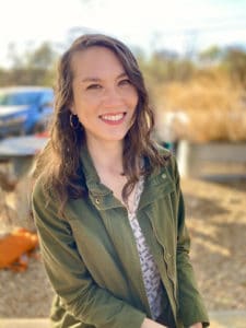 A photograph of Miranda Nadeau, PhD, Licensed Psychologist and owner of Panorama Therapy. She is smiling and making eye contact with the viewer. She wears a green utility jacket over a black and white patterned top and gold earrings. There is a golden cast to the photo. Behind her, out of focus, are picnic tables and a blue car.